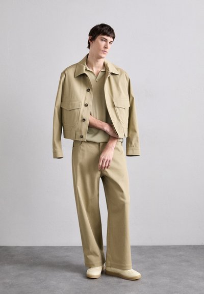 Beige bomulljacka med två framfickor, tillsammans med en matchande beige kortärmad skjorta och byxor med vida ben, bärs med krämfärgade sneakers.