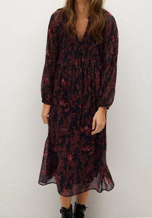 Femme portant une robe midi noire à motifs floraux rouges, avec des manches longues transparentes, et des bottines noires, debout devant un fond clair uni.