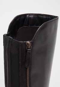 Gros plan d'une botte en cuir noir avec une fermeture éclair latérale et un panneau élastique sous la fermeture éclair pour un enfilage plus facile et un ajustement flexible.