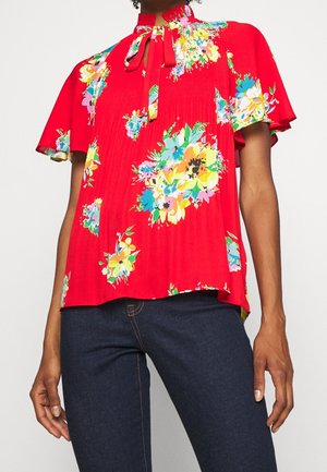 Red short-sleeve blouse with multicolored floral print, high neck tie, worn with dark blue jeans.
