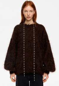 Joven con cabello rojo que lleva un suéter peludo marrón oscuro y un collar largo de plata con cuentas, posando frente a un fondo blanco liso.