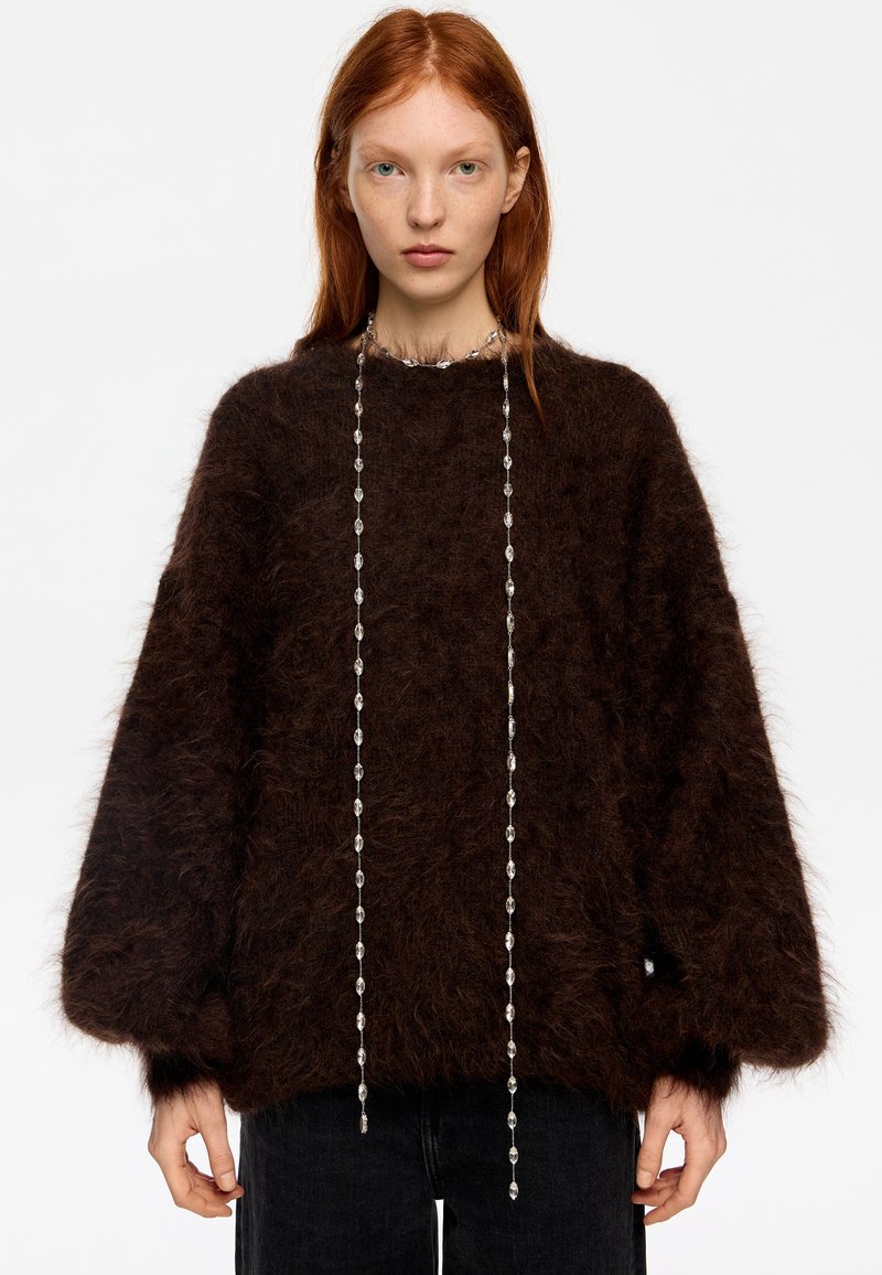 Joven con cabello rojo que lleva un suéter peludo marrón oscuro y un collar largo de plata con cuentas, posando frente a un fondo blanco liso.