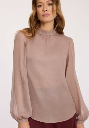 Kobieta w przezroczystej, długosleewej bluzce w kolorze brudnego różu z wysokim dekoltem i subtelnym połyskiem, zestawiona z ciemną spódnicą lub spodniami.