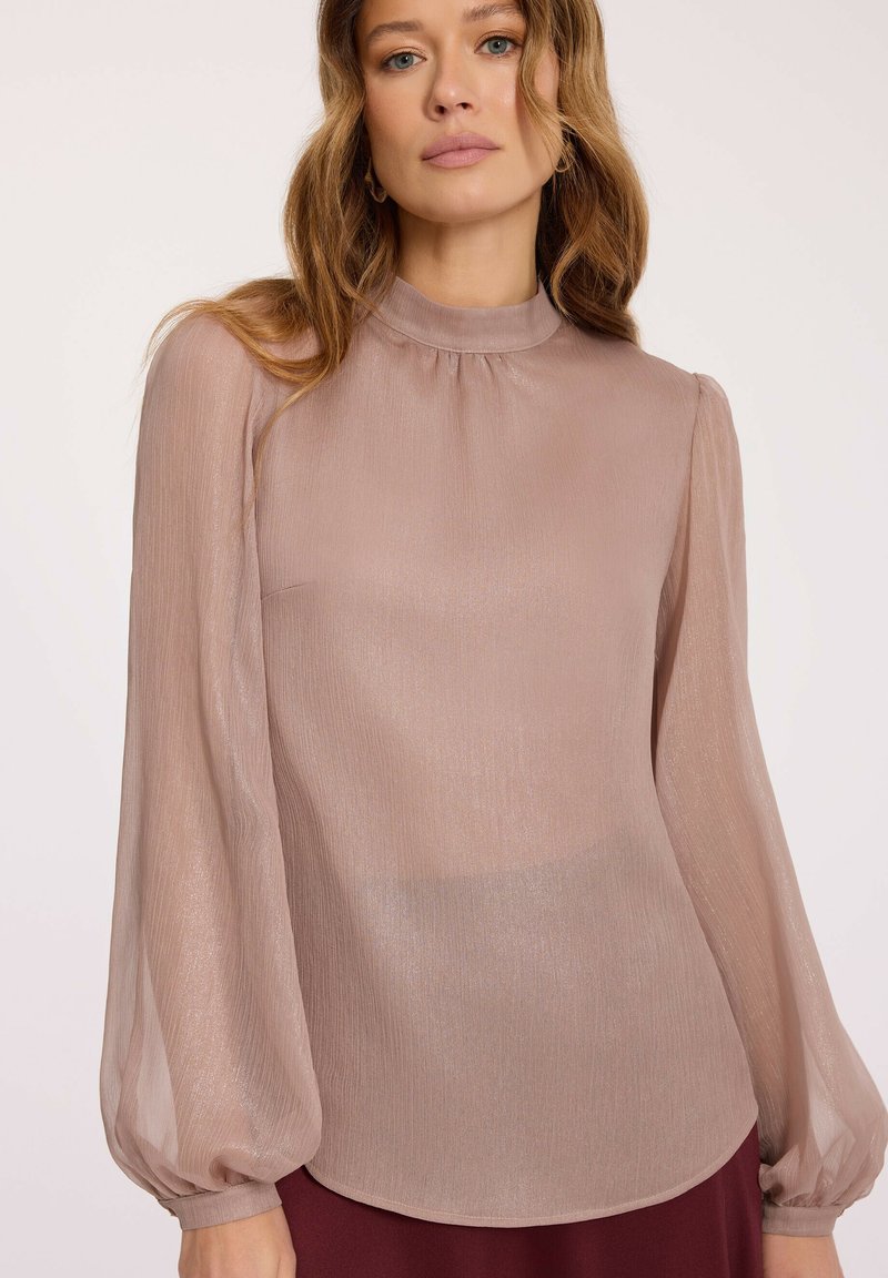 Kobieta w przezroczystej, długosleewej bluzce w kolorze brudnego różu z wysokim dekoltem i subtelnym połyskiem, zestawiona z ciemną spódnicą lub spodniami.