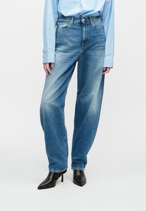 Žena v bledomodrých rovno strihaných džínsoch, čiernych špicatých podpätkových čižmách a svetlomodrej pruhovanej dlhopristej blúzke.