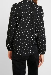 Blusa nera con pois bianchi, scollatura arricciata e polsini elasticizzati. Design casual con una leggera caduta e orlo arrotondato.