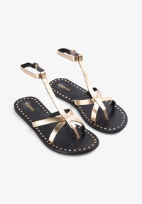 Gold metallic strappy sandals with a black sole, featuring stud accents and a toe post design. Brand name "SCALPERS" is printed on the insole.