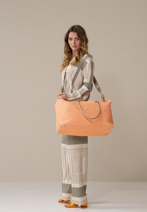 Grote, koraalkleurige tote bag met gestructureerde weefsel, draaghandvaten en gouden letters. Draagt bij een gestreept beige en grijs outfit.