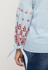 Chemise rayée bleu clair et blanche avec broderie florale rouge sur les manches, présentant un accent de poignet noué et une texture lisse.