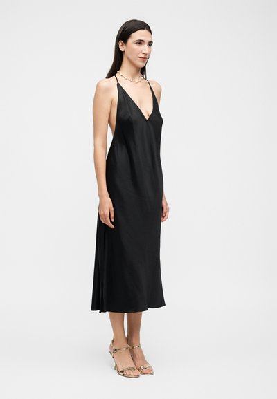 Γυναίκα με μακριά σκούρα μαλλιά που φοράει μια μαύρη σατέν μίντι φόρεμα χωρίς μανίκια με βαθύ V-neck και χρυσά ψηλοτάκουνα πέδιλα, στέκεται σε απλό φόντο.