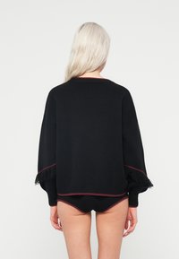 Μαύρο πουλόβερ με χαλαρή εφαρμογή, διακοσμημένο με κόκκινη επένδυση και φούντες στα μανίκια, συνδυασμένο με σλιπ μαύρης ραφής.