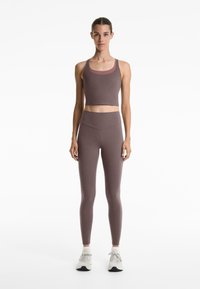 Brun sport-bh och matchande högmidjade leggings i ett mjukt, stretchigt material. Modellen har på sig ljusa sneakers och står framför en enkel bakgrund.