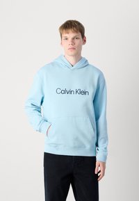 Svetlo modrá mikina s kapucňou vyrobená z jemného materiálu, s prednou klokankovou vreckom a logom "Calvin Klein" v tmavej farbe na hrudi.
