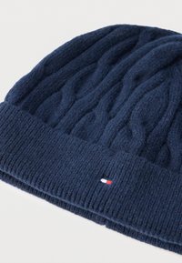 Σκούφος πλεκτός σκούρου μπλε χρώματος με σχέδια σε σχήμα καλωδίου και ριμπ μανσέτα. Δι�αθέτει μια μικρή ετικέτα με λογότυπο σε κόκκινο, λευκό και μπλε στο πλάι.