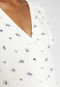Vestido floral branco com padrões azuis e verdes, apresentando um decote em V, mangas curtas e botões redondos. Textura subtil em riscas verticais.