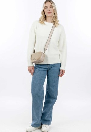 Weißer flauschiger Pullover, hellblaue Jeans und eine beigefarbene Umhängetasche mit einem Fellverschluss. Model steht vor einem weißen Hintergrund.
