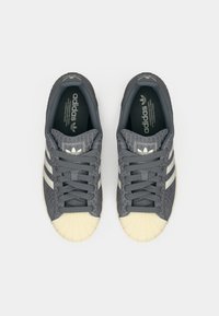 Γκρι αθλητικό παπούτσι με υφασμένη επάνω επιφάνεια, κρεμ ελαστική προστατευτική μύτη και τρεις λευκές ρίγες. Διαθέτει γκρι κορδόνια και εκτυπωμένη εσωτερική ετικέτα.