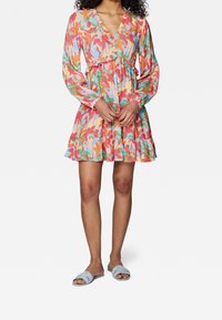 Robe florale colorée avec un décolleté en V, des manches longues et un ourlet à volants. Portée avec des sandales plates bleu clair. Tissu lisse avec un motif vibrant.
