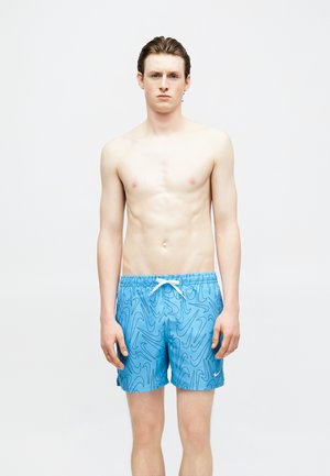 Νέος άντρας με κοντά καστανά μαλλιά, ντυμένος με μπλε μαγιό σορτς που έχουν μαύρα μοτίβα Nike, στέκεται μπροστά σε ένα μονόχρωμο λευκό φόντο.