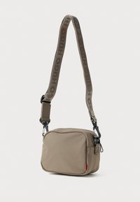 Brun stofcrossbody tas med lynlås, justerbar rem med logo og en lille rød detalje på siden.