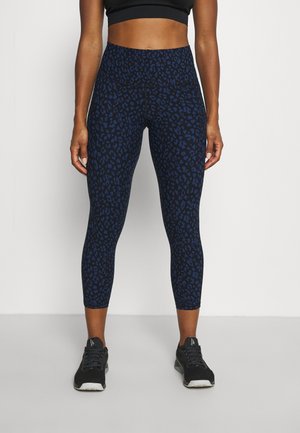 Vrouw die hooggetailleerde cropped legging met een zwarte en donkblauwe luipaardprint en zwarte sportschoenen draagt, staand tegen een witte achtergrond.