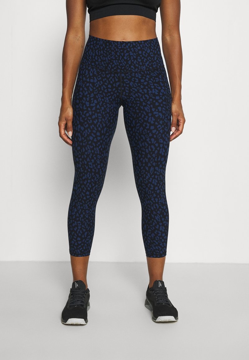 Femme portant des leggings courts à taille haute avec un imprimé léopard noir et bleu foncé et des chaussures de sport noires, debout devant un fond blanc.