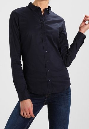 Personne portant une chemise noire ajustée à manches longues boutonnée et un jean bleu foncé, posant avec un bras plié et l'autre main reposant sur la cuisse.