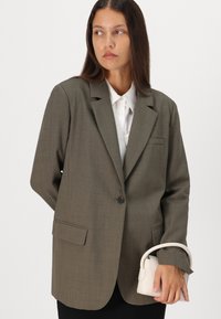 Blazer vert olive au design structuré, doté d'une fermeture à un bouton et de deux poches avant. Tient un petit sac à main crème.