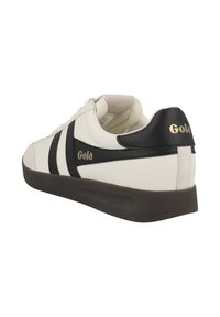 Zapatilla de cuero blanca con rayas y talón negros. Presenta un collar acolchado, puntera redonda y suela de goma marrón. Logo "Gola" en dorado.