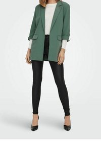 Vihreä ylisuuri bleiseri, jossa on käärityt hihat, kaksi etutaskua, yhdistetty mustiin tiukkoihin housuihin ja korkokenkiin. Sileä rakenne.