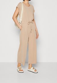 Beige jumpsuit met korte mouwen en een getailleerde taille, voorzien van wijde pijpen. Gecombineerd met witte sandalen en een lichtgekleurde crossbodytas.