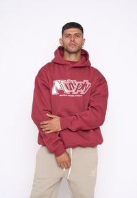 Felpa cappuccio bordeaux con logo grafico bianco, caratterizzata da una vestibilità comoda e polsini a costine. Indossata con pantaloni della tuta beige chiaro.