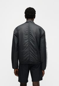 Zwarte bomberjack met elastische zoom en manchetten, voorzien van een gladde, glanzende textuur. Twee witte strepen lopen langs de mouwen.