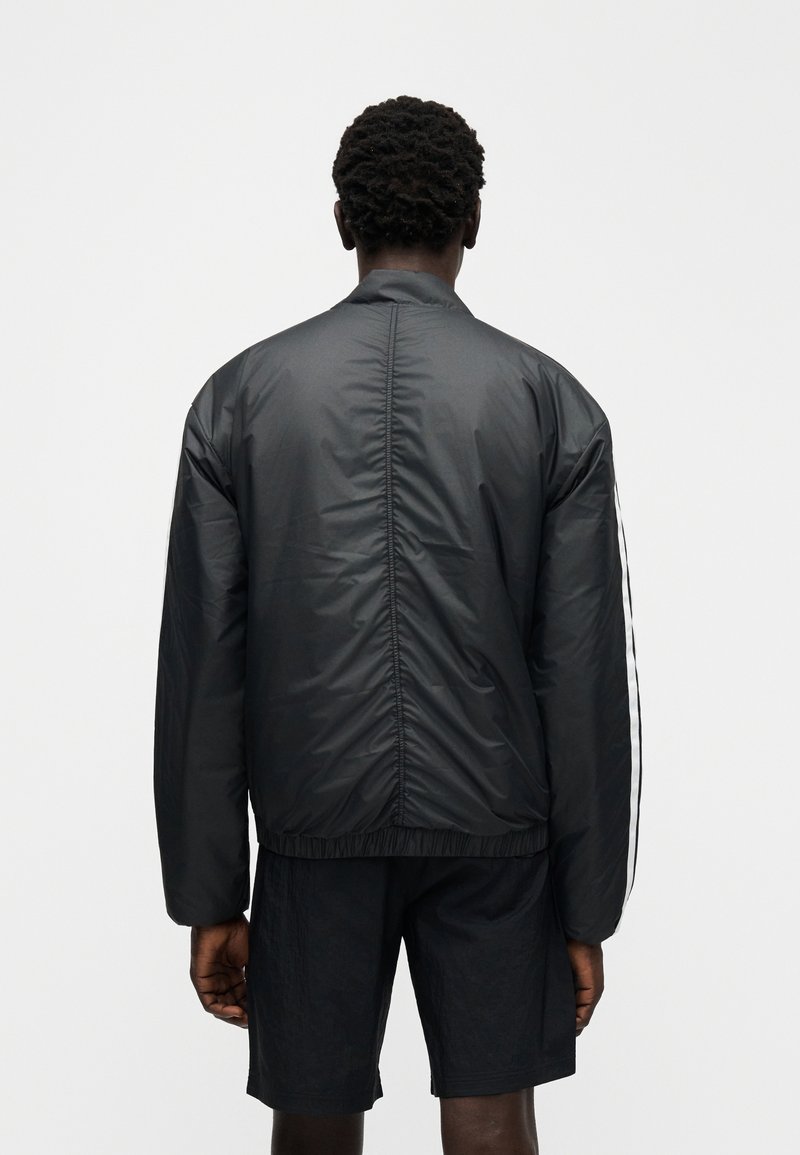 Schwarze Bomberjacke mit elastischem Saum und Bündchen, die eine glatte, glänzende Textur aufweist. Zwei weiße Streifen verlaufen entlang der Ärmel.