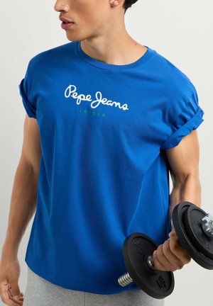 Mann in blauem Pepe Jeans T-Shirt hebt eine schwarze Hantel mit hochgekrempelten Ärmeln vor einfarbigem Hintergrund.