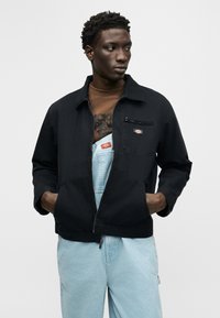 Νέος με σκούρα σγουρά μαλλιά, που φοράει μαύρο μπουφάν με φερμουάρ πάνω από καφέ πουκάμισο και ανοιχτόχρωμα τζιν παντελόνια εργασίας, κοιτάζοντας προς το πλάι.