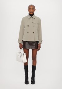 Giacca beige doppiopetto con bottoni scuri, dolcevita nera, mini gonna in pelle marrone, stivali neri alti fino al ginocchio e borsa bianca.