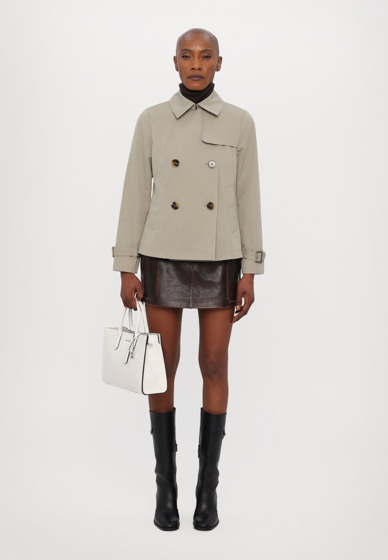 Giacca beige doppiopetto con bottoni scuri, dolcevita nera, mini gonna in pelle marrone, stivali neri alti fino al ginocchio e borsa bianca.