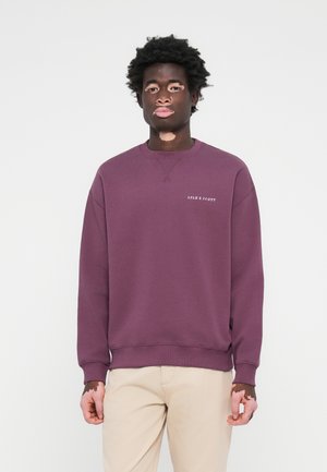 Lila sweatshirt med rund hals och ribbade ärmslut, med en liten vit logotyp på vänster sida. Bärs med ljusbeiga byxor.