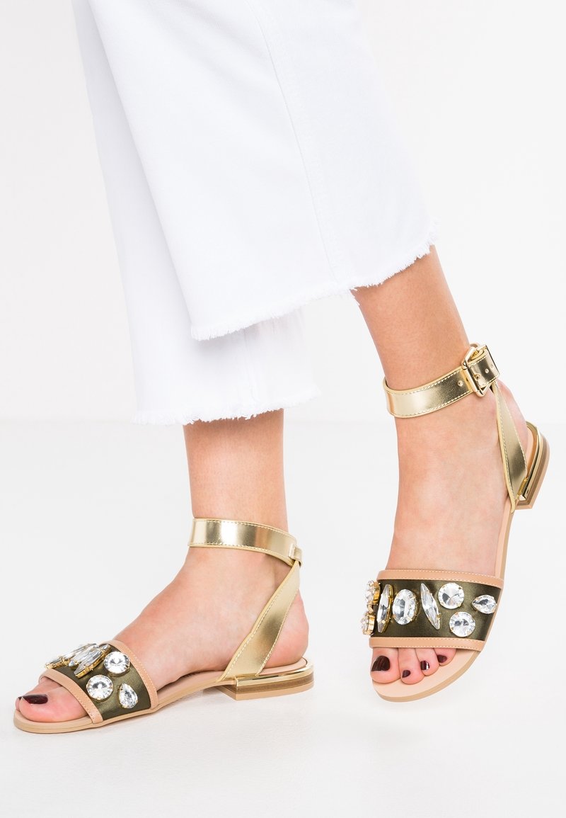 Pieds portant des sandales dorées à bout ouvert avec de grosses pierres décoratives transparentes et des brides à la cheville, associées à un pantalon blanc court avec des ourlets effilochés.