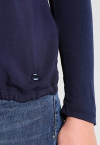 Camiseta de manga larga en color navy con dobladillo redondeado, que presenta una cinturilla elástica fruncida y un botón decorativo negro en la parte inferior.
