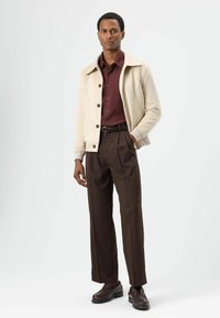 Beige leren jas met een kraag, gedragen over een donkerrood overhemd en bruine hoge taillebroek. Zwarte riem en donkere schoenen maken de look compleet.