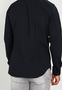 Mężczyzna w czarnej koszuli z długim rękawem i jasnoszarych spodniach, pokazany od tyłu z prawą ręką swobodnie opadającą wzdłuż ciała.