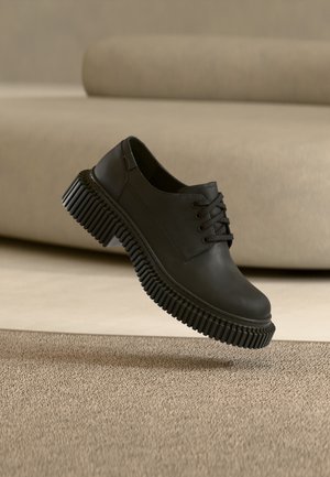 Zapato negro de vestir con cordones y suela gruesa acanalada parece levitar sobre una alfombra beige texturizada y un fondo neutro.