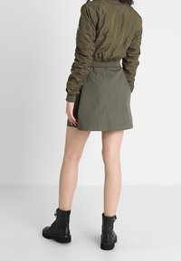 Osoba w oliwkowym bomberze i pasującej spódnicy, w czarnych sznurowanych botkach za kostkę, stojąca na tle jednolitego białego tła.