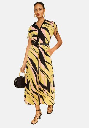 Žena má na sebe midi šaty so žltým, ružovým a čiernym abstraktným vzorom s čiernym opaskom, drží čiernu okrúhlu kabelku a má na sebe čierne sandále na podpätku so remienkami.