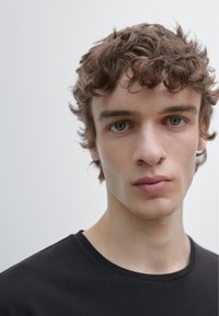 Sort bomulds-T-shirt med rund halsudskæring, båret af en ung mand med bølget, mellemlangt hår og neutral baggrund.