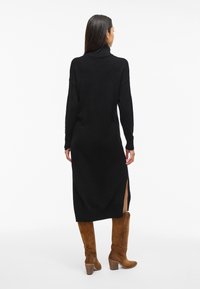 Svart stickad tröjklänning med hög krage, långa ärmar, sidospjäll och avslappnad passform, kombinerad med bruna mockastövlar som når knäna.