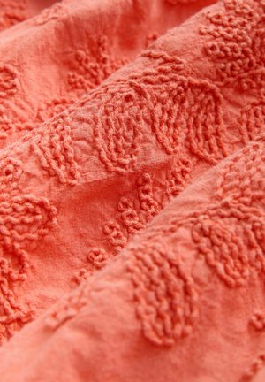 Korallfärgat tyg med texturerad broderi och upphöjda mönster, som visar detaljerat stygnarbete och en mjuk, vävd yta.