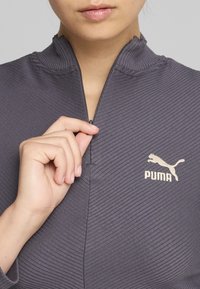 Sivi, rebrasti pulover s četvrtim patentnim zatvaračem i visokim ovratnikom. Ima zlatni Puma logo na prsima. Ruka drži povlačnik patentnog zatvarača.